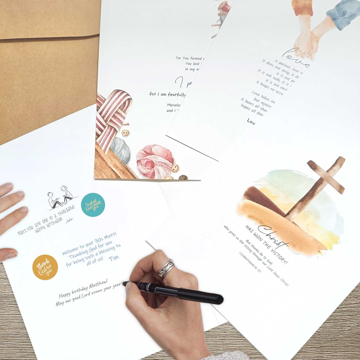 Several printed greeting cards and a kraft envelope are arranged on a wooden surface, with a hand holding a black pen writing on one card. The cards feature watercolor illustrations, including  a wooden cross over a landscape.
