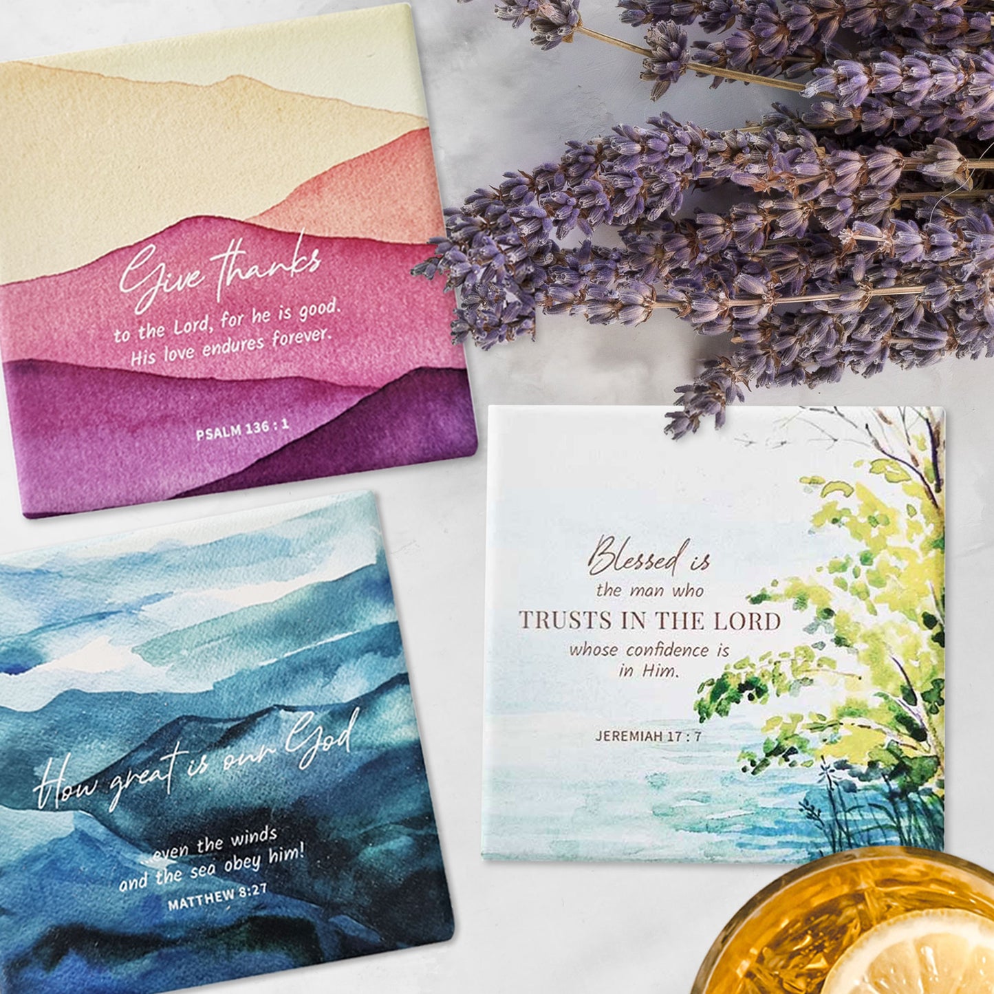 Three square ceramic coasters with Bible verses flat lay on a marble top with a glass of drink and lavendar flowers.