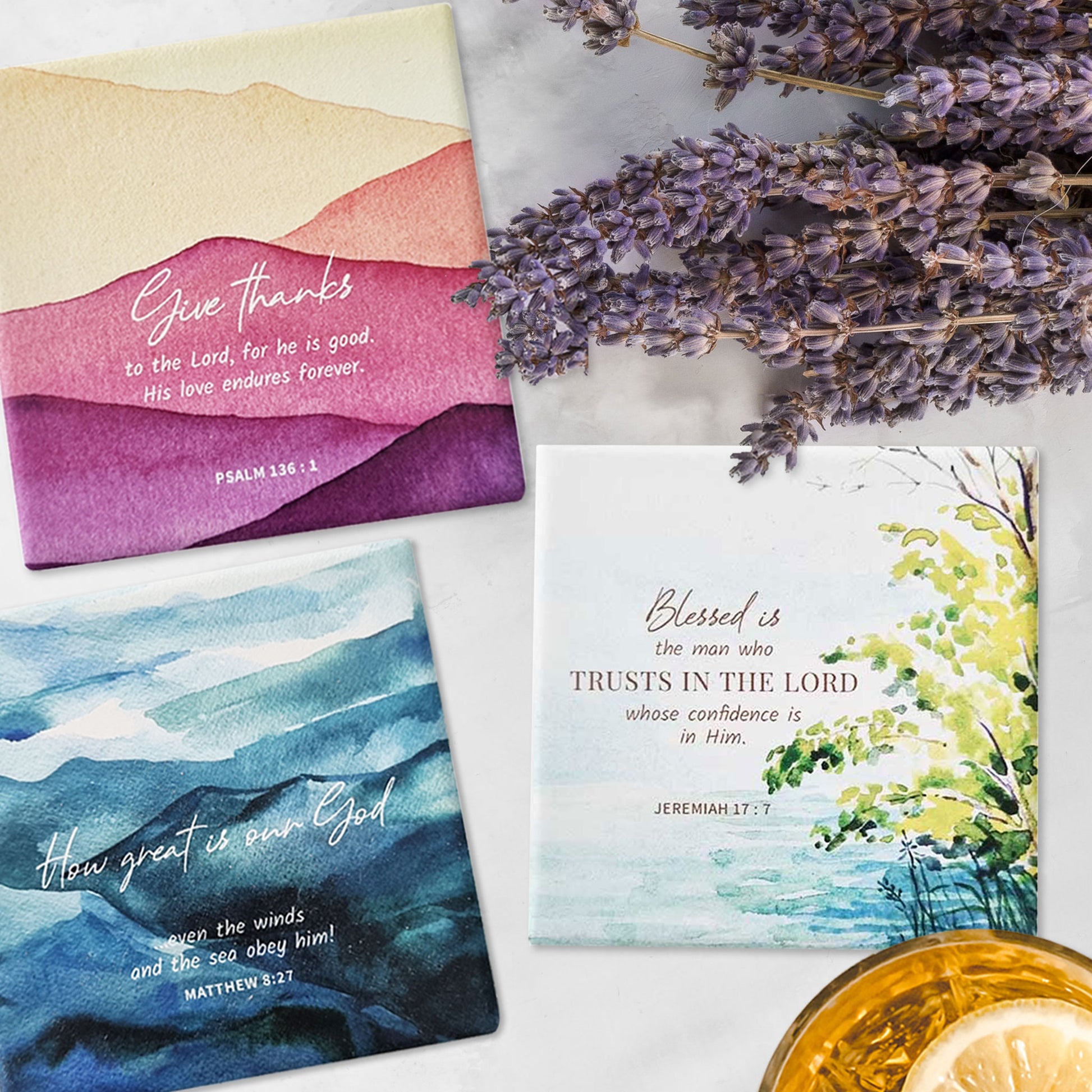 Three square ceramic coasters with Bible verses flat lay on a marble top with a glass of drink and lavendar flowers.