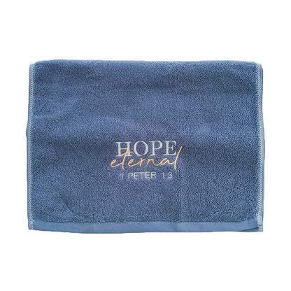 Folded blue cotton towel with embroidered text 'Hope eternal 1 Peter 1:3' on a white background