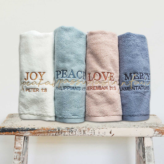Four rolled cotton towels with embroidered text referencing Christian Bible verses on a wooden stool against a light background