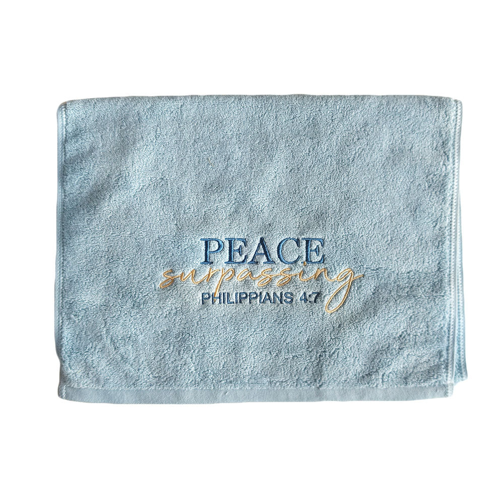 Folded turquoise cotton towel with embroidered text 'Peace surpassing Philippians 4:7' on a white background