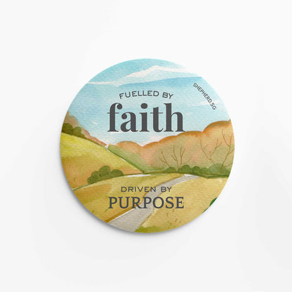 Round pin badge with text "Fuelled by faith driven by purpose" and watercolor road and sky design on a white background