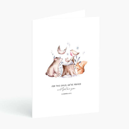 A white greeting card standing upright against a light gray background, featuring a watercolor illustration of woodland animals including a bear, fox, bird, and crescent moon, with the text 'FOR THIS CHILD, WE'VE PRAYED and God has given'.