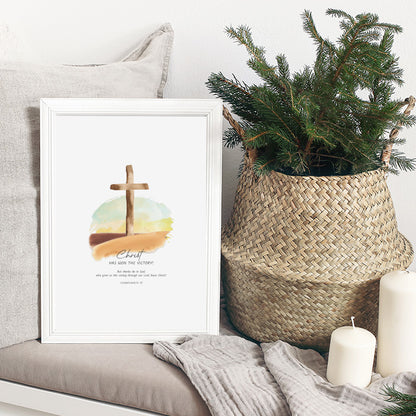 A framed art print featuring a watercolor cross over a sunset with the text 'Christ has won the victory', styled on a sofa next to a woven basket with greenery and candles.