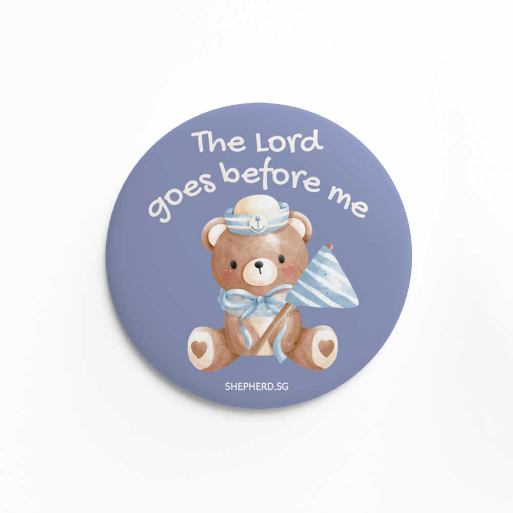 Button pin badge with teddy bear holding a flag design and 'The Lord goes before me' text on a white background