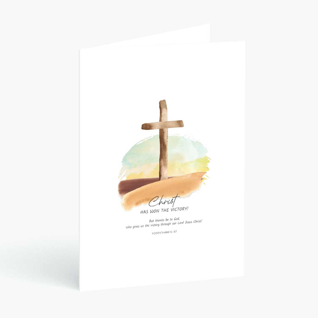 A white greeting card standing upright, featuring a watercolor illustration of a wooden cross over a soft blue, yellow, and brown landscape. Below the artwork, the text reads, 'Christ has won the victory!' 